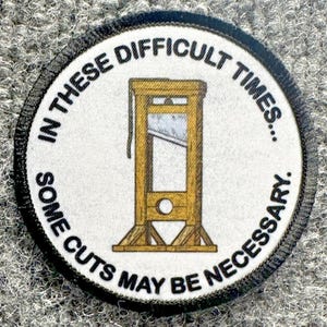 May include: A circular embroidered patch with a white background and a black stitched border. The center features a brown illustration of a guillotine. The text reads: IN THESE DIFFICULT TIMES... SOME CUTS MAY BE NECESSARY in black capital letters.