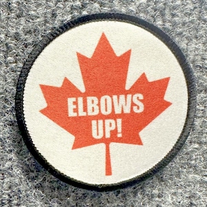 May include: A circular embroidered patch with a white background and a black border. Centered on the patch is a large orange maple leaf graphic. The text ELBOWS UP! is printed in white capital letters across the center of the maple leaf.