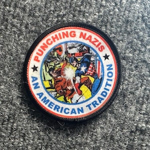May include: A circular embroidered patch featuring a comic book illustration of a superhero punching a figure. The border is white with red text reading PUNCHING NAZIS and blue text reading AN AMERICAN TRADITION, separated by two black stars.