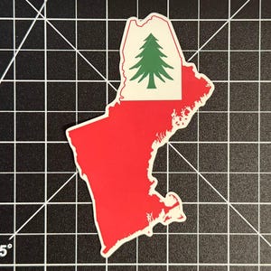 May include: A die-cut sticker shaped like the state of Maine, featuring a solid red background with a white rectangular inset at the top containing a green pine tree graphic. The sticker has a thin white border and is displayed on a black grid mat.