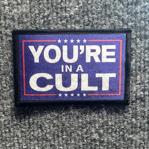 May include: A rectangular patch with a dark blue background and a thin red border. The text YOU'RE IN A CULT is printed in large white capital letters. Five small white stars are aligned horizontally above and below the central text.