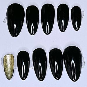 May include: A set of black, glossy, oval-shaped press-on nails, with one gold glitter nail. The nails are arranged on a white card, set against a dark, textured background.