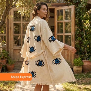 May include: Cream-colored robe with a repeating pattern of blue eye designs. The robe has a belt, wide sleeves, and a fringed hem. The background includes a wooden door and greenery.