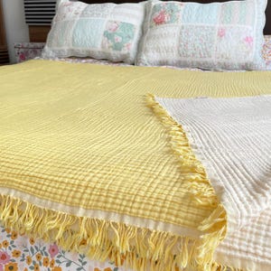 May include: A yellow and white fringed blanket is draped over a bed. The blanket is made of a textured fabric. Two decorative pillows with floral patterns are in the background. The bedspread has a floral pattern.