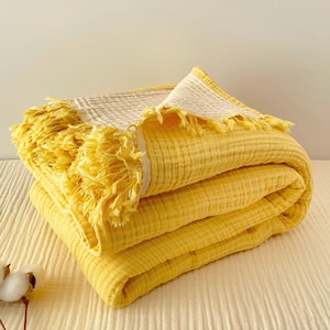 Peut inclure: Couverture jaune pliée avec une bordure à franges. La couverture est faite d'un matériau doux et texturé et est pliée avec soin. La couche supérieure a une section blanche. La couverture est empilée sur une surface crème.