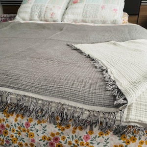 May include: A two-tone grey and white blanket with fringing. The blanket is on a bed with floral patterned sheets and pillows. The blanket has a soft, lightweight texture. The fringe is grey and white.