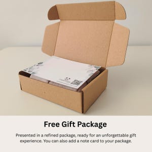 May include: A brown cardboard shipping box with an open lid containing a white rectangular card with a botanical leaf border design, a small panda logo, and a QR code printed on the surface, resting atop a folded light pink fabric item.