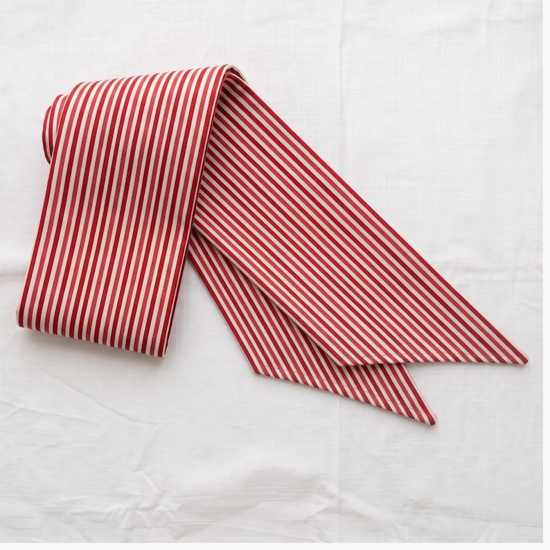 May include: A long, narrow fabric sash or hair ribbon featuring a repeating pattern of vertical red and white stripes. The accessory is folded over itself, displaying a consistent striped design across its entire length and pointed ends.