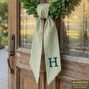 May include: A long fabric ribbon with a green and white vertical striped pattern, tied in a knot at the top and hanging down. The right side of the ribbon features a large, dark green capital letter H embroidered on the fabric surface.