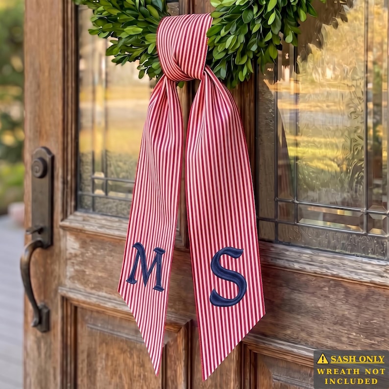 May include: A red and white vertically striped fabric sash featuring the embroidered navy blue letters M and S on the two hanging tails. The sash is tied in a knot at the top and is displayed hanging from a green leaf wreath on a wooden door.