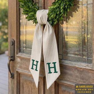 May include: A beige and white striped fabric sash featuring a dark green embroidered letter H on each of the two hanging tails. The sash is tied around a green boxwood wreath. Text on the image reads: SASH ONLY WREATH NOT INCLUDED.