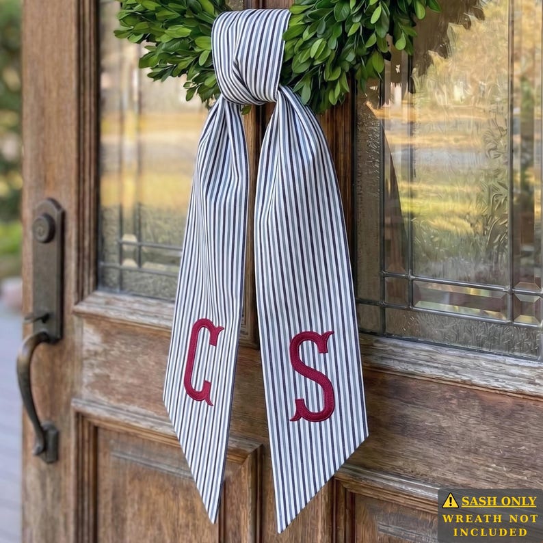 May include: A long fabric ribbon with a black and white vertical pinstripe pattern, tied in a knot at the top. The two hanging ends feature the embroidered red letters C and S. The ribbon is attached to a green wreath hanging on a wooden door.