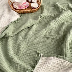 May include: A light green and off-white reversible blanket with a fringed edge. A woven tray holds a teacup, saucer, and a small candle. The blanket has a textured, crinkled appearance, suggesting a soft, breathable fabric.