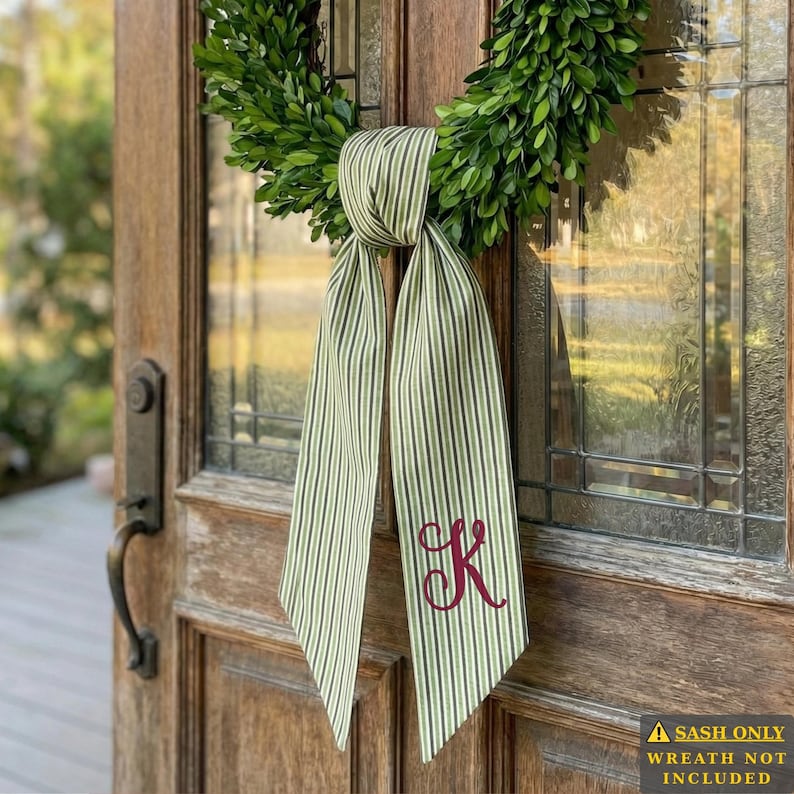 A close-up view of a fabric sash with vertical green and white stripes, tied to a lush green boxwood wreath. Near the bottom corner of the right-hand tail, there is a beautifully embroidered decorative letter 'K' in a rich burgundy thread. The sash hangs in front of an old-fashioned wooden door with leaded glass panels.