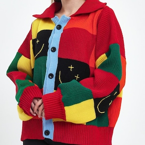 Puede incluir: Un cárdigan de patchwork colorido con cuello y cierre de botones. El suéter presenta una mezcla de paneles rojos, amarillos, verdes y negros, con una tapeta de botones azul claro y botones negros. Las mangas y el dobladillo son rojos.