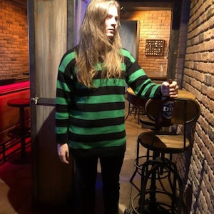 May include: A green and black striped sweater is worn with black pants and gray sneakers. The person is holding a bottle of beer. The background includes a brick wall, wooden door, and a bar stool.