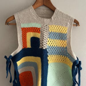 Può includere: Un gilet all'uncinetto fatto a mano con un design patchwork. Il gilet presenta una base beige neutra con quadrati colorati di arancione, giallo, blu e verde. I lacci blu scuro sono sui lati. Il gilet è appeso a una gruccia di legno.