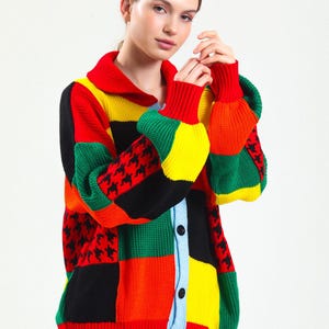 Può includere: Un cardigan patchwork colorato con collo e chiusura a bottoni. Il maglione presenta un mix di pannelli rossi, gialli, verdi, neri e arancioni, con dettagli a pied-de-poule. Le maniche e il colletto sono rosse.