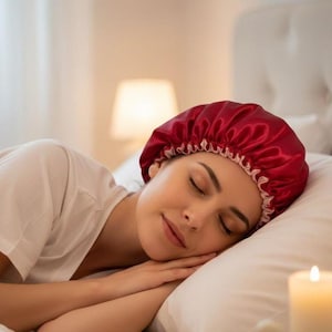 May include: A person wearing a burgundy satin sleep cap with a white trim, resting on a white pillow. The cap is a deep red color and appears soft and smooth. The person is in a relaxed position.