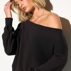 Könnte beinhalten: Schwarzes Off-Shoulder-Sweatshirt mit langen Ärmeln. Das Oberteil ist aus einem weichen Material gefertigt und hat eine lockere Passform. Eine goldene Halskette ist sichtbar. Das Model trägt eine blaue Jeans. Der Hintergrund ist weiß.
