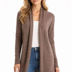 May include: A long, brown cardigan with a shawl collar. The cardigan is worn over a white top and blue jeans. The sleeves are long and fitted, and the cardigan falls to mid-thigh. The overall style is casual and comfortable.
