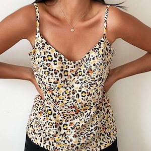 Può includere: Canotta senza maniche con scollo a V e spalline sottili. La parte superiore presenta una stampa leopardata a macchie nere, marroni, arancioni e gialle su sfondo crema. Il capo è realizzato in tessuto leggero.