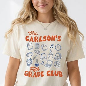 May include: A cream-colored short-sleeve t-shirt featuring the orange text Mrs. CARLSON'S Fifth GRADE CLUB. Below the text are blue line-art icons of school supplies including notebooks, a calculator, a globe, a backpack, and a laptop computer.
