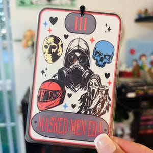 May include: A rectangular air freshener with a black string. The design features illustrations of a gas mask, a hockey mask, a skull, a motorcycle helmet, and a figure in a ghost mask holding a knife. The text "MASKED MEN ERA" is displayed.