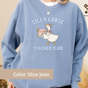 May include: A blue jean colored crewneck sweatshirt featuring a graphic of a white goose wearing a pink bow and flower, carrying a stack of books. The text reads SILLY GOOSE TEACHER CLUB in light blue letters. The design is centered on the chest.