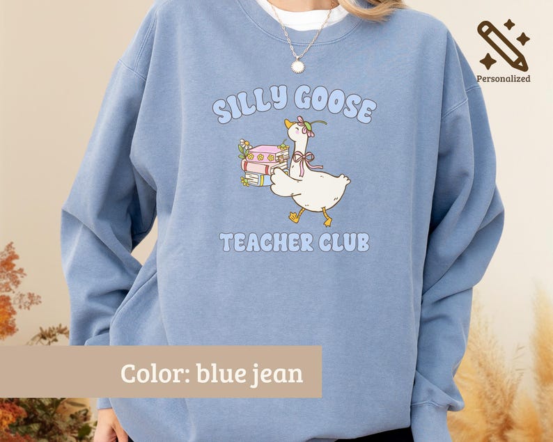 May include: A blue jean colored crewneck sweatshirt featuring a graphic of a white goose wearing a pink bow and flower, carrying a stack of books. The text reads SILLY GOOSE TEACHER CLUB in light blue letters. The design is centered on the chest.