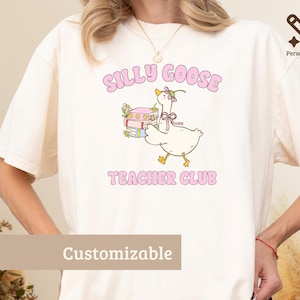 May include: A cream-colored t-shirt featuring a graphic of a white goose wearing a flower crown and a pink bow, carrying a stack of books. The text above the goose reads SILLY GOOSE and the text below reads TEACHER CLUB in pink letters.
