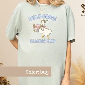 May include: A bay-colored t-shirt featuring a graphic of a white goose wearing a small hat and bow, standing next to a stack of books. The text "SILLY GOOSE TEACHER CLUB" is printed in light blue letters above and below the goose illustration.