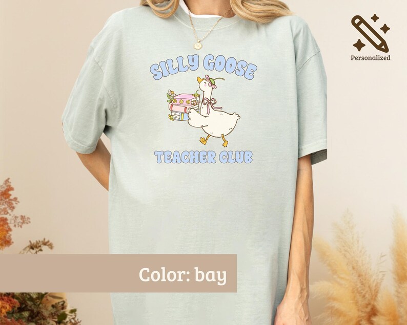 May include: A bay-colored t-shirt featuring a graphic of a white goose wearing a small hat and bow, standing next to a stack of books. The text "SILLY GOOSE TEACHER CLUB" is printed in light blue letters above and below the goose illustration.