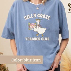 May include: A blue jean colored t-shirt featuring a graphic of a white goose wearing a pink bow and flower crown, carrying a stack of books. The text reads SILLY GOOSE TEACHER CLUB in white letters. The shirt has short sleeves and a crew neckline.