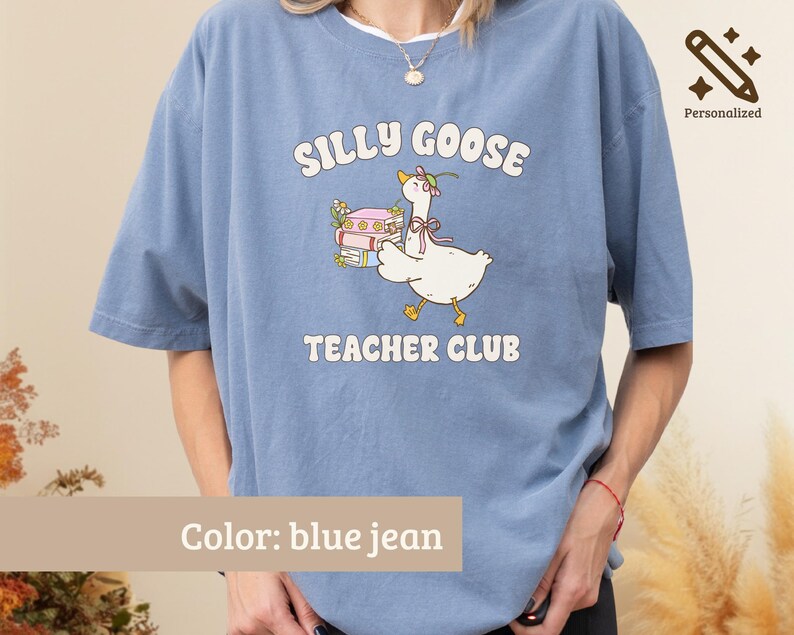 May include: A blue jean colored t-shirt featuring a graphic of a white goose wearing a pink bow and flower crown, carrying a stack of books. The text reads SILLY GOOSE TEACHER CLUB in white letters. The shirt has short sleeves and a crew neckline.