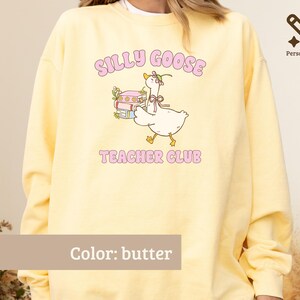 May include: A butter yellow crewneck sweatshirt featuring a graphic of a goose wearing a bonnet and carrying a stack of books. The text "SILLY GOOSE" appears above the goose and "TEACHER CLUB" below, both in pink lettering. The design includes small floral accents.