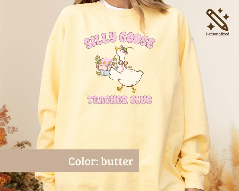 May include: A butter yellow crewneck sweatshirt featuring a graphic of a goose wearing a bonnet and carrying a stack of books. The text "SILLY GOOSE" appears above the goose and "TEACHER CLUB" below, both in pink lettering. The design includes small floral accents.