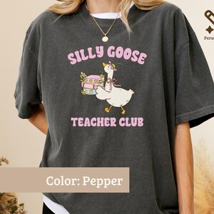May include: A pepper-colored t-shirt featuring a graphic of a white goose wearing a flower crown and a pink ribbon, standing next to a stack of books. The text reads SILLY GOOSE TEACHER CLUB in pink letters above and below the goose illustration.