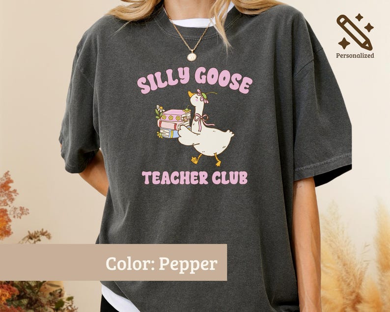 May include: A pepper-colored t-shirt featuring a graphic of a white goose wearing a flower crown and a pink ribbon, standing next to a stack of books. The text reads SILLY GOOSE TEACHER CLUB in pink letters above and below the goose illustration.