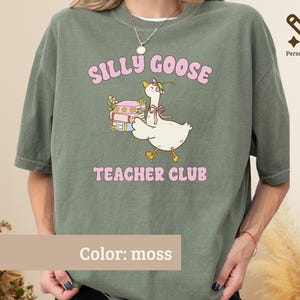 May include: A moss green t-shirt featuring a graphic of a white goose wearing a flower crown and a bow, standing next to a stack of books. The text "SILLY GOOSE" appears above the goose and "TEACHER CLUB" appears below in pink lettering.