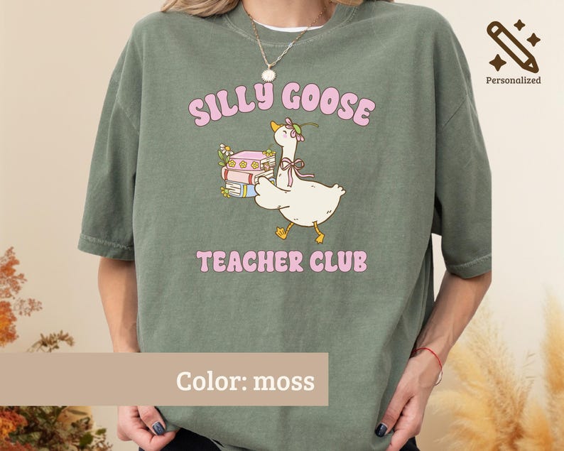 May include: A moss green t-shirt featuring a graphic of a white goose wearing a flower crown and a bow, standing next to a stack of books. The text "SILLY GOOSE" appears above the goose and "TEACHER CLUB" appears below in pink lettering.