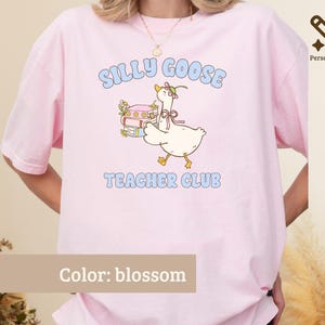 May include: A blossom pink t-shirt featuring a graphic of a white goose wearing a flower crown and a pink ribbon, standing next to a stack of books. The text reads SILLY GOOSE TEACHER CLUB in light blue letters above and below the illustration.
