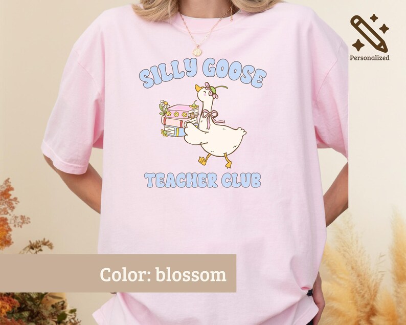 May include: A blossom pink t-shirt featuring a graphic of a white goose wearing a flower crown and a pink ribbon, standing next to a stack of books. The text reads SILLY GOOSE TEACHER CLUB in light blue letters above and below the illustration.