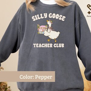 May include: A pepper-colored crewneck sweatshirt featuring a graphic of a white goose wearing a floral bonnet and a pink ribbon, carrying a stack of colorful books. The text reads SILLY GOOSE TEACHER CLUB in a cream-colored, rounded font.
