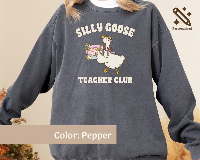 May include: A pepper-colored crewneck sweatshirt featuring a graphic of a white goose wearing a floral bonnet and a pink ribbon, carrying a stack of colorful books. The text reads SILLY GOOSE TEACHER CLUB in a cream-colored, rounded font.