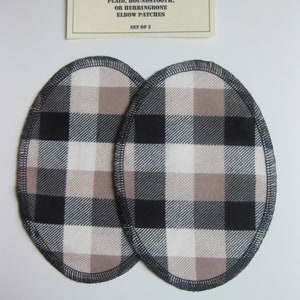 Puede incluir: Dos parches de codo ovalados con un patrón de cuadros negros, blancos y beige. Los bordes están acabados con una puntada de sobrehilado negra. Una etiqueta crema en la parte superior dice "CRAFT T'S PLAID, HOUNDSTOOTH, OR HERRINGBONE ELBOW PATCHES SET OF 2."