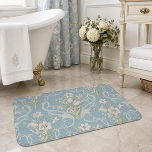 May include: A rectangular bath mat featuring a light blue background with a repeating pattern of white floral bouquets and swirling vine motifs. The mat has rounded corners and a soft, flat-weave texture, positioned on a light-colored tile floor.