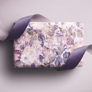 Op de afbeelding: Een rechthoekige geschenkdoos verpakt in bloemenpatroonpapier met witte, paarse en groene tinten. Een paars satijnen lint is om de doos gedrapeerd en creëert een decoratief accent. De achtergrond is zacht lavendelkleurig.