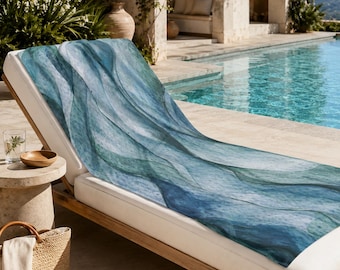 May include: A rectangular beach towel featuring an abstract watercolour pattern of flowing, layered waves in shades of deep navy, teal, and soft seafoam green, draped over a white cushioned lounge chair near a swimming pool.