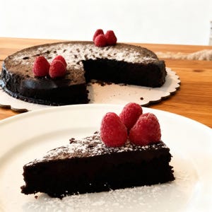 Op de afbeelding: Een stuk donkere chocoladetaart, bestrooid met poedersuiker en versierd met verse frambozen, op een wit bord. Een hele chocoladetaart, op dezelfde manier versierd, staat op de achtergrond.